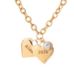 Gold necklace with heart-shaped pendants engraved with 'Alex' and '2026', featuring a clear gemstone on a white background.