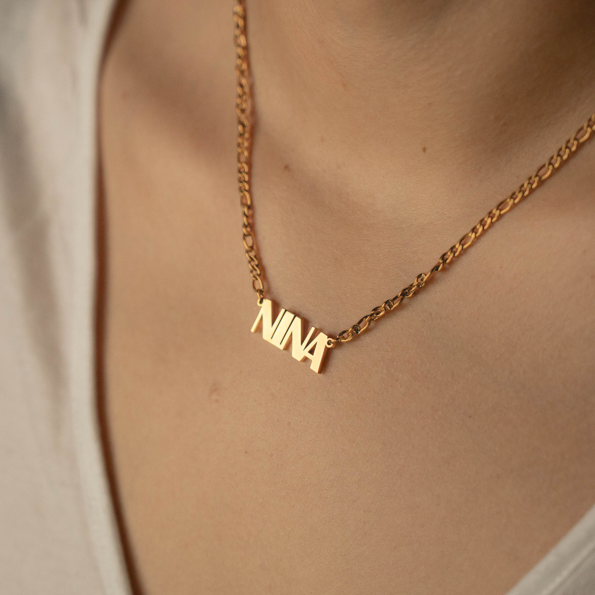 A close-up of a person wearing a gold chain necklace with a custom nameplate pendant spelling 'NINA'.