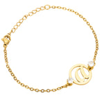 A gold-colored bracelet featuring a single initial letter surrounded by two small clear stones, displayed on a white background.